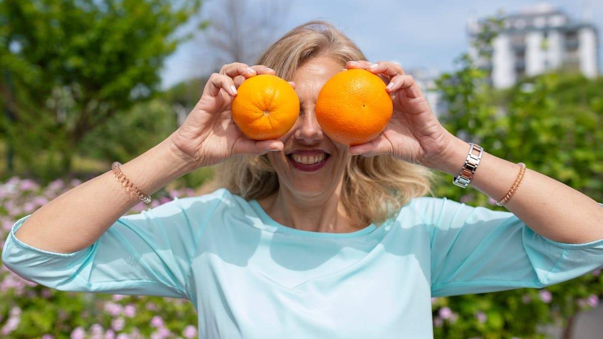 A smiling woman playfully holding oranges in a sunny park setting.